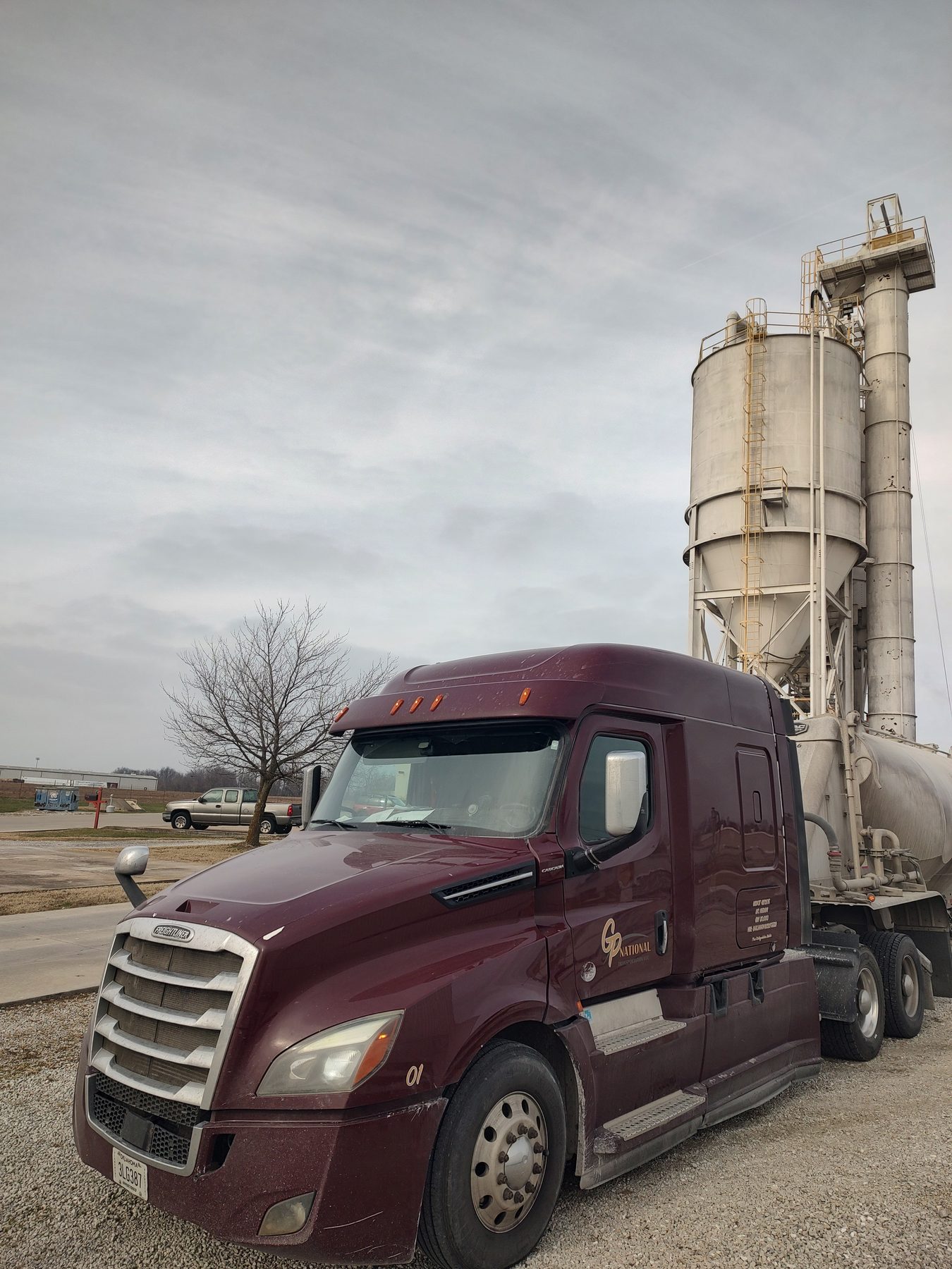 GP National maroon Freightliner Cascadia parked in front of storage silos under a cloudy sky